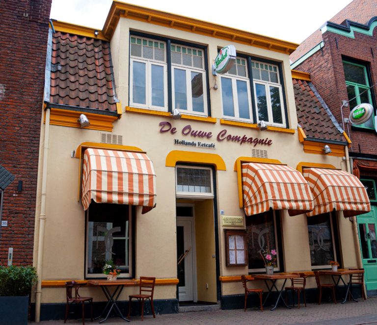De gele gevel van De Ouwe Compagnie met gestreepte zonneschermen en terras
